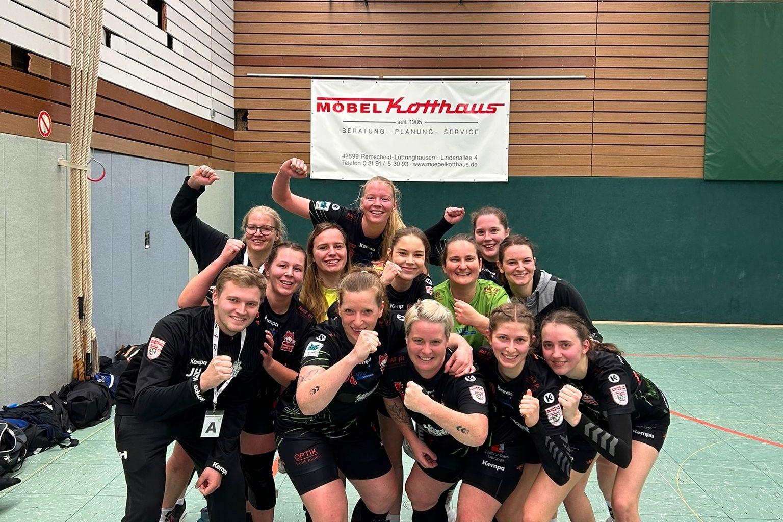 Gruppensiegpose von Handballspielerinnen in einem Sportgymnasium, mit Punktestand und Werbebanner im Hintergrund.