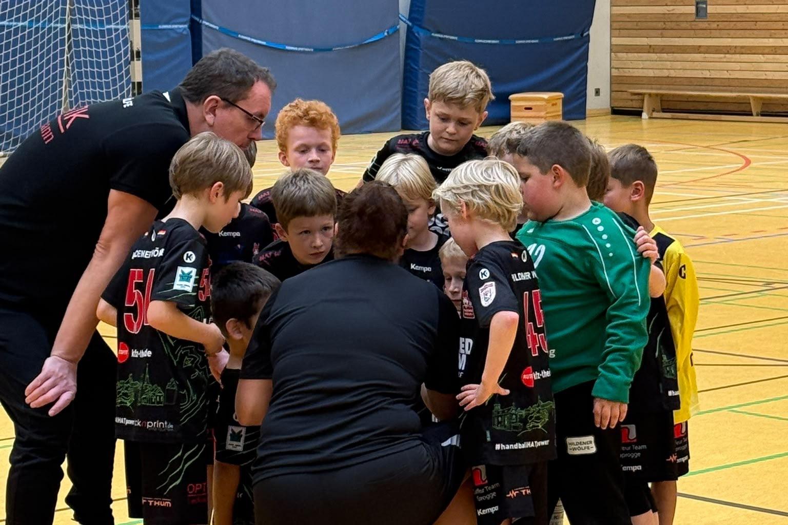 Gruppe von Kindern in Sportkleidung huddelt sich um Trainer in einer Sporthalle.