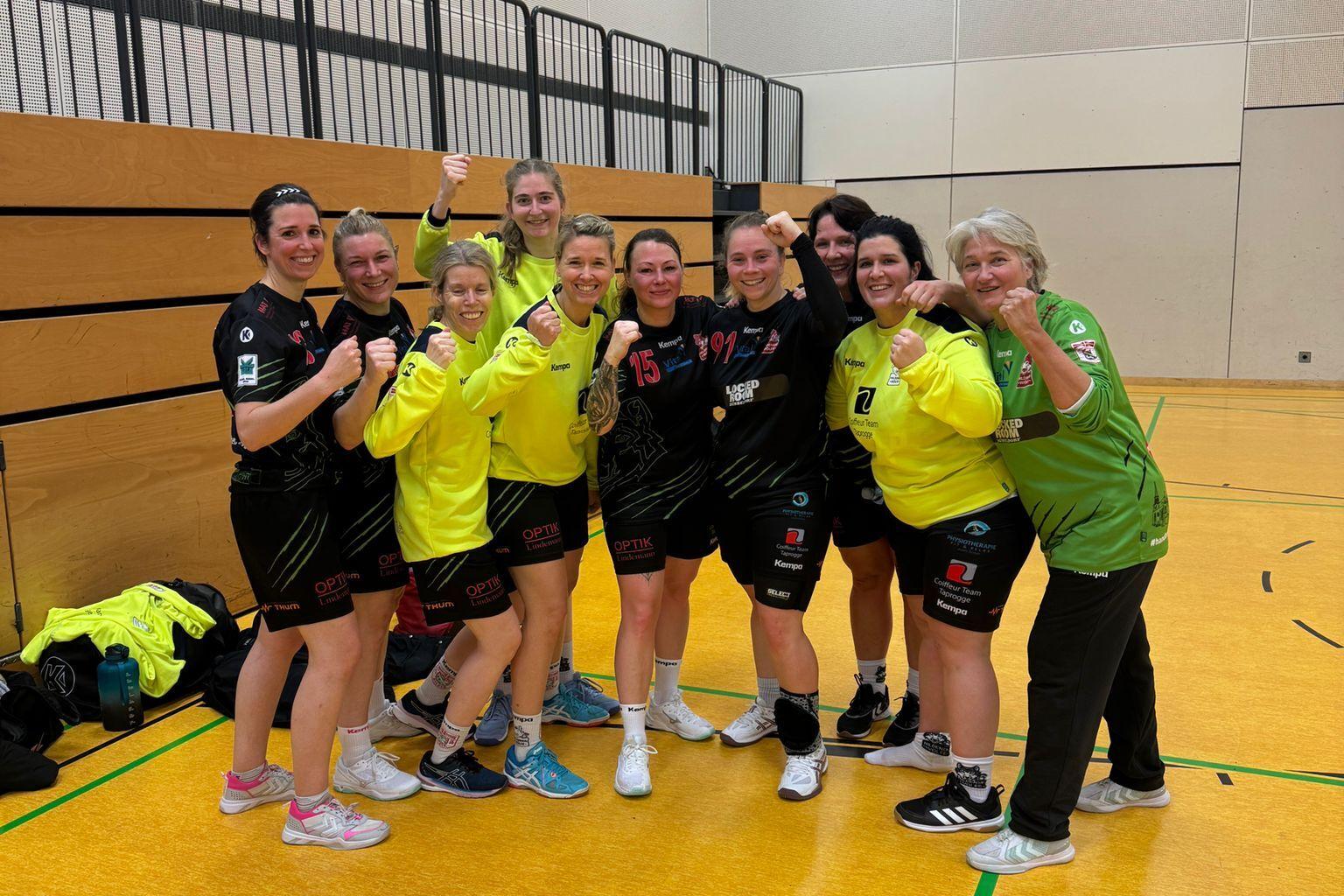 Gruppenfoto von zehn Handballspielerinnen in Sportkleidung, die im Trainingsraum posieren und zum Sieg zeigen.