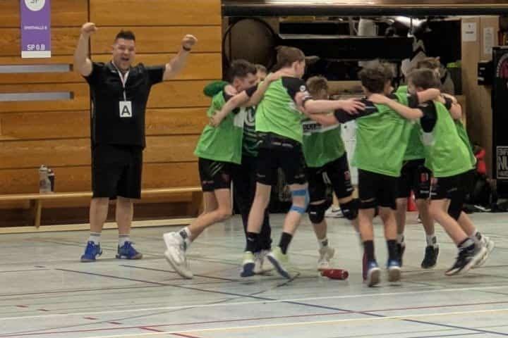 Gruppe von Kindern in grünen Trikots feiert zusammen, während ein Trainer mit ausgestreckten Armen beobachtet.