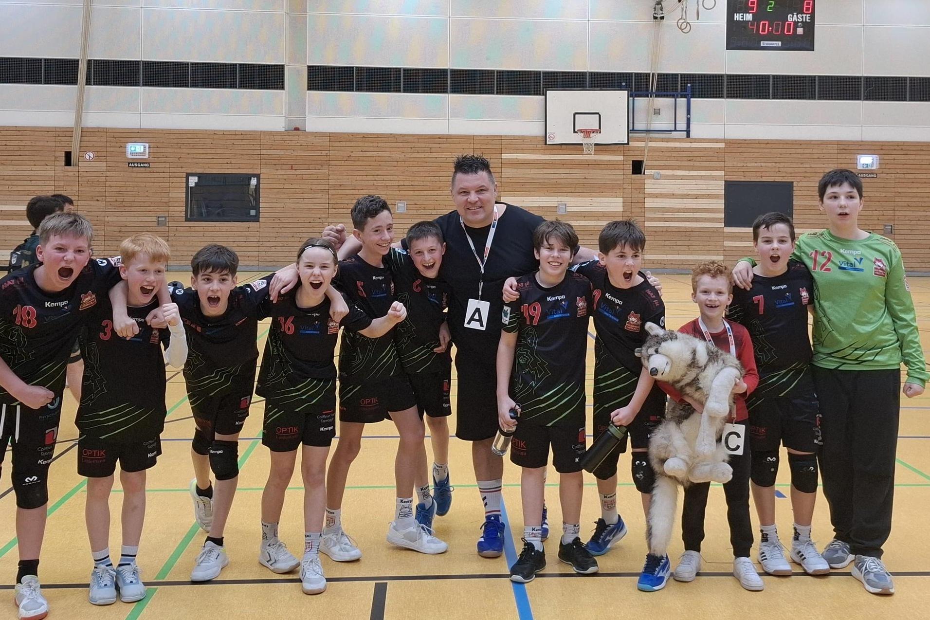 Junge Handballmannschaft posiert jubilierend mit Trainer in Sporthalle, einige halten ein Pl&uuml;schtier.