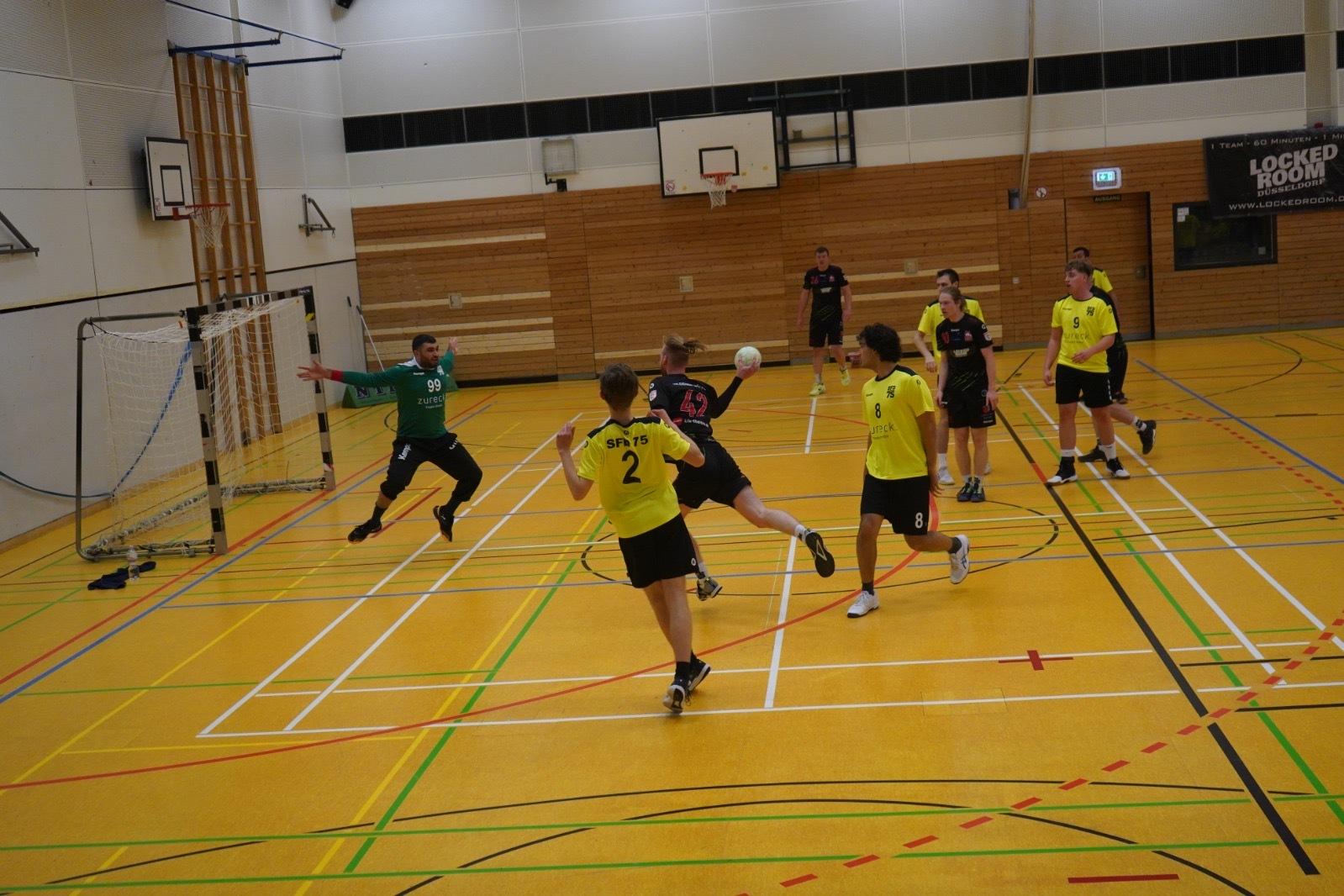 Zwei Handballmannschaften in gelben und schwarzen Trikots spielen in einer Sporthalle. Der Torwart springt zur Abwehr.