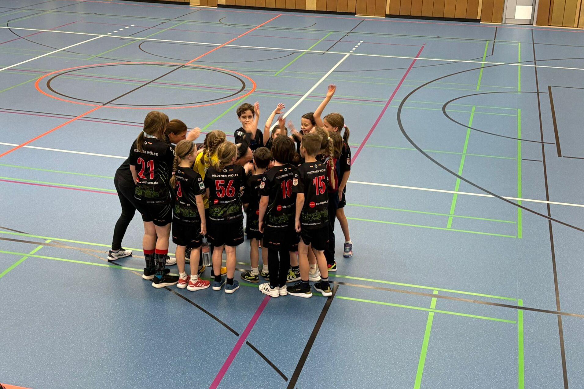 Mädchenmannschaft in schwarzer Sportkleidung bildet einen Kreis auf einem Sportplatz mit markierten Linien.
