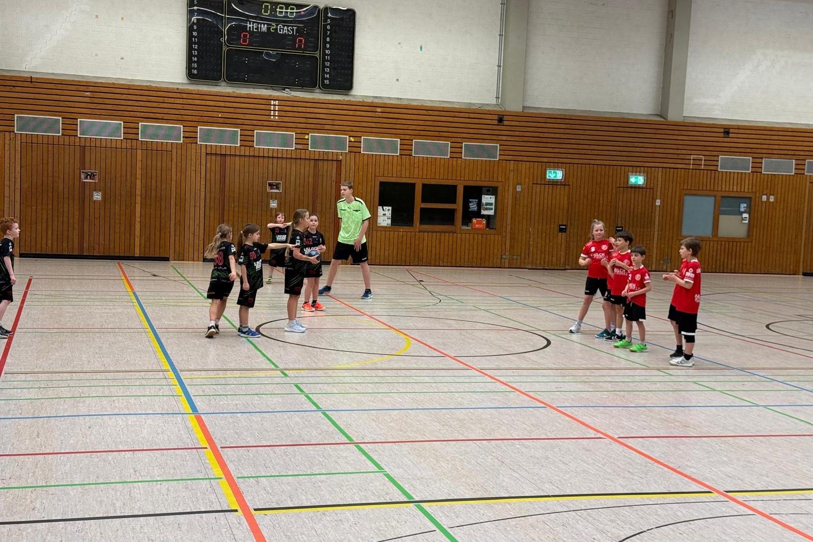 Zwei Gruppen von Kindern in Sportkleidung spielen Handball in einer hellen Halle mit markierten Linien auf dem Boden.