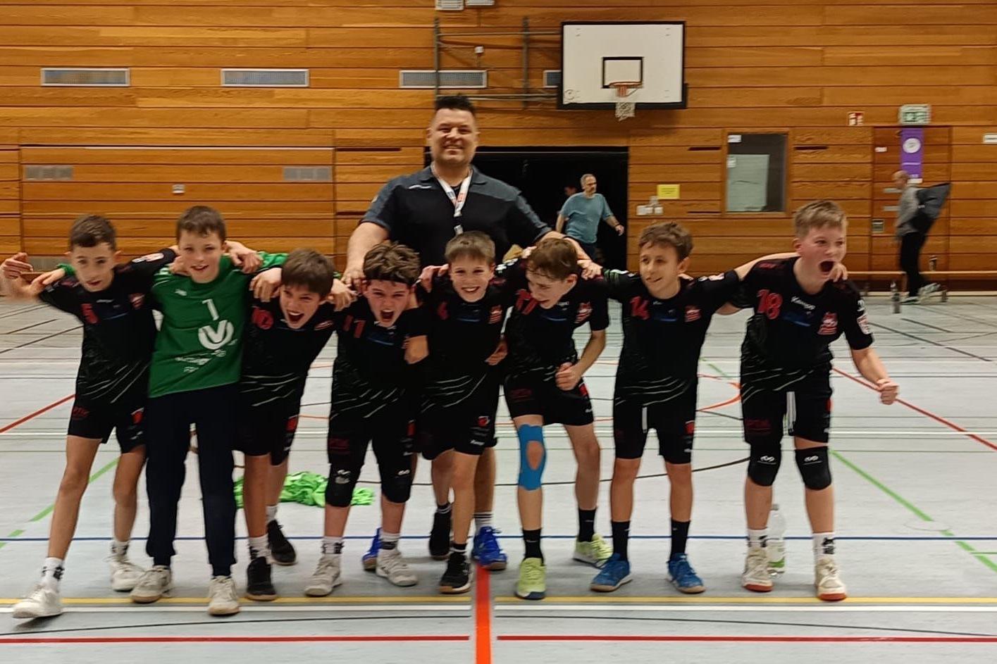 Gruppe junger Handballspieler in schwarz-roten Trikots posiert lachend mit Trainer in Sporthalle.