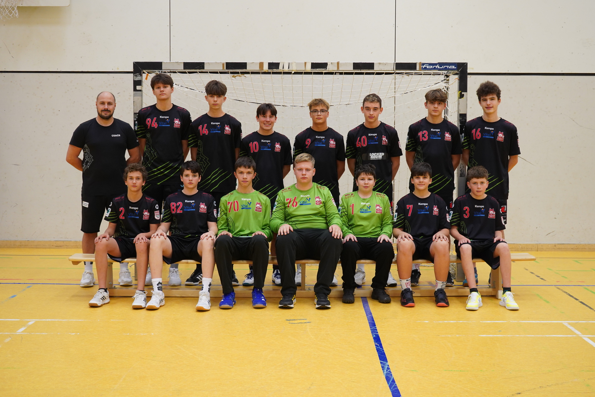 Gruppenfoto einer Jugend-Handballmannschaft mit Trainer, alle tragen einheitliche Trikots in einer Sporthalle.