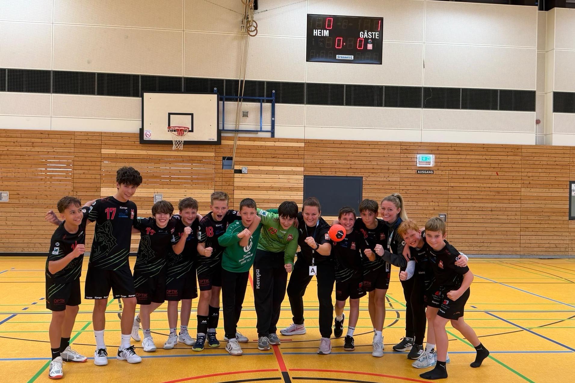 Gruppe von Handballspielern und Trainerin posieren l&auml;chelnd in sportlicher Kleidung auf dem Spielfeld einer Sporthalle.