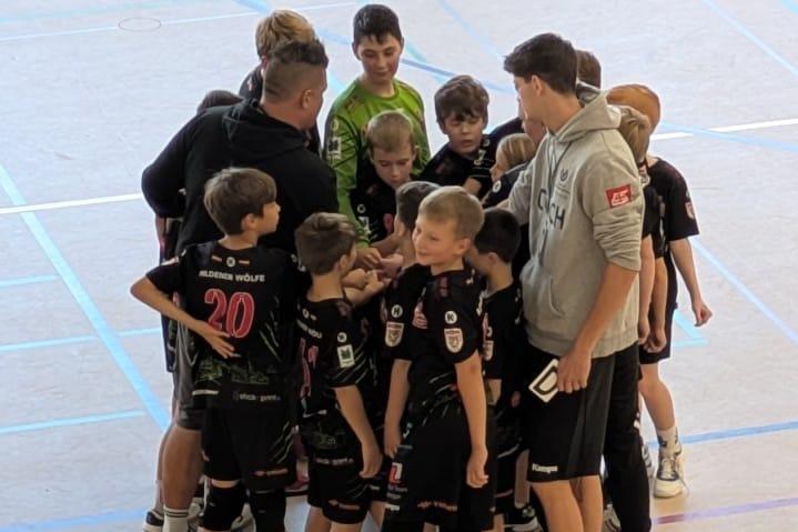 Gruppe von Jungen in Sportkleidung um einen Trainer versammelt, in einer Halle mit bunten Linien auf dem Boden.