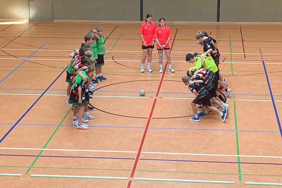 Zwei Teams in Sportkleidung stehen vor einem Ball auf dem Hallenboden, bereit für das Spiel. Die Uhr zeigt 15 Minuten.