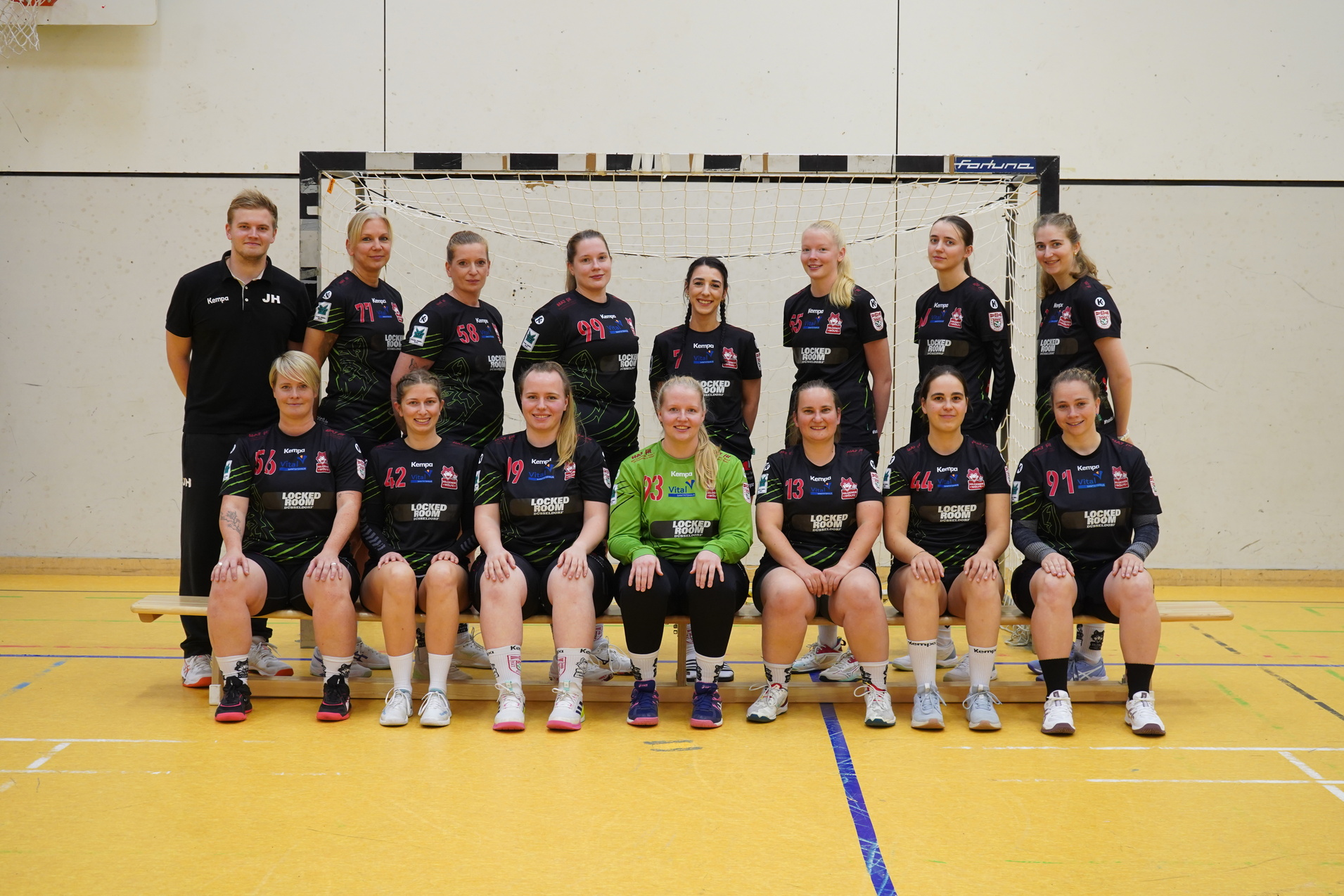 Handballmannschaft mit Trainer pose auf dem Feld vor dem Tor, alle in schwarzen Trikots mit Sponsorenlogos.