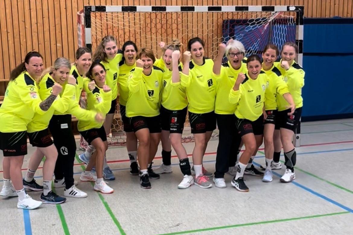 Gruppe von Handballspielerinnen in leuchtend gelben Trikots posiert freudig vor einem Handballtor in einer Sporthalle.