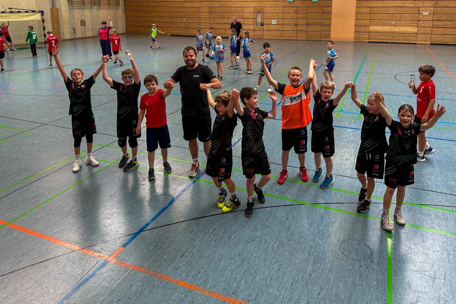 Gruppe von Kindern in Sportkleidung, die jubelnd in einer Halle stehen, umgeben von anderen Spielern im Hintergrund.