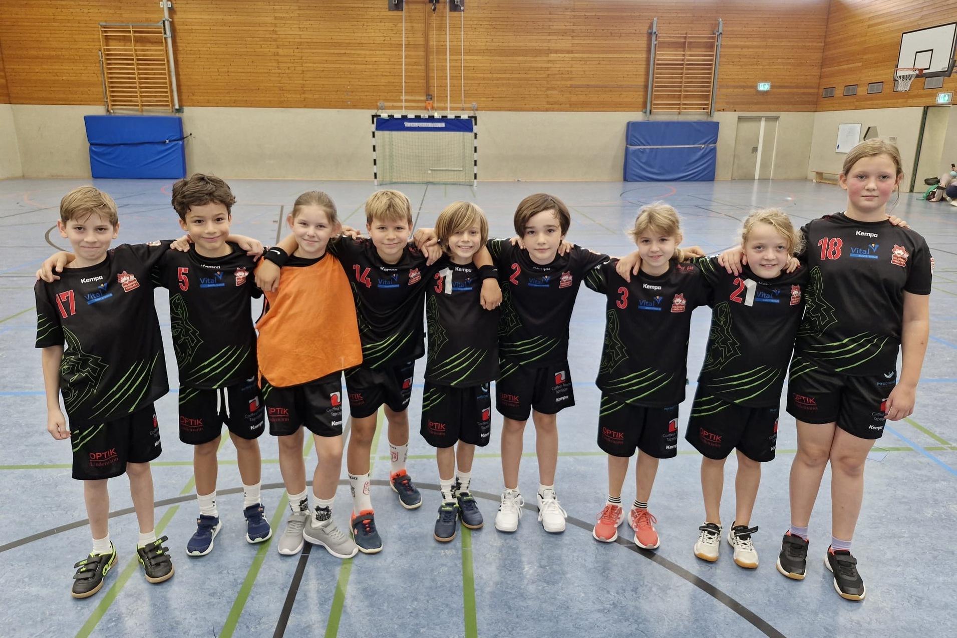 Gruppe von zehn Kindern in Sportkleidung, lächelnd, auf einem Handballfeld in einer Sporthalle.