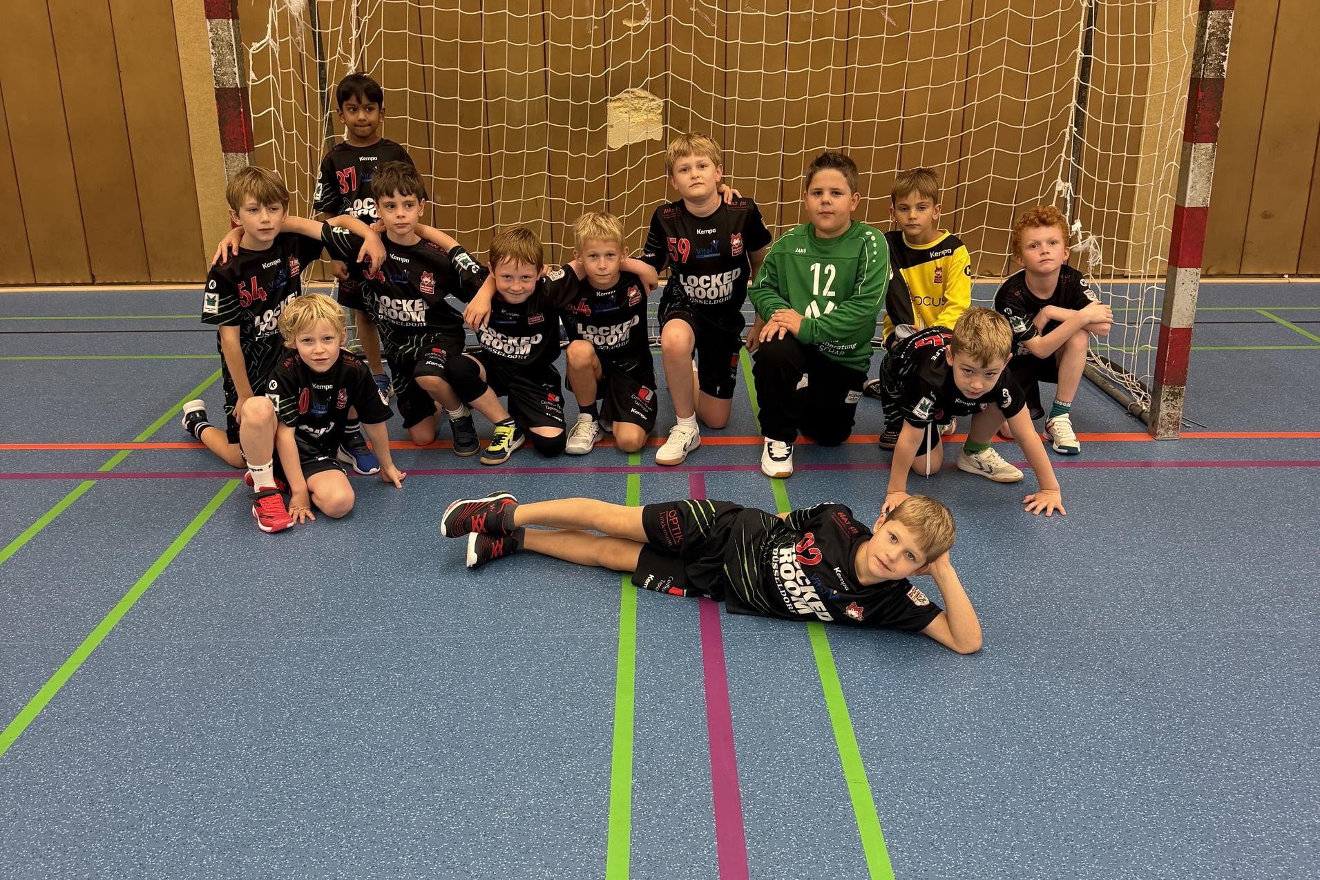 Gruppe von zwölf Kindern in Sportkleidung, posierend vor einem Handballtor in einer Halle.