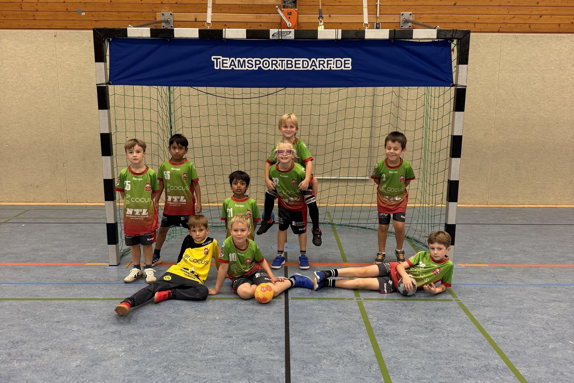 Gruppe von Kindern in Sportkleidung vor einem Handballtor in einer Turnhalle, einige sitzen, andere stehen.