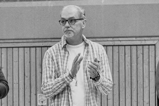 Mann mit Brille und Karotenhemd applaudiert in einer Halle; Holzverkleidung im Hintergrund.