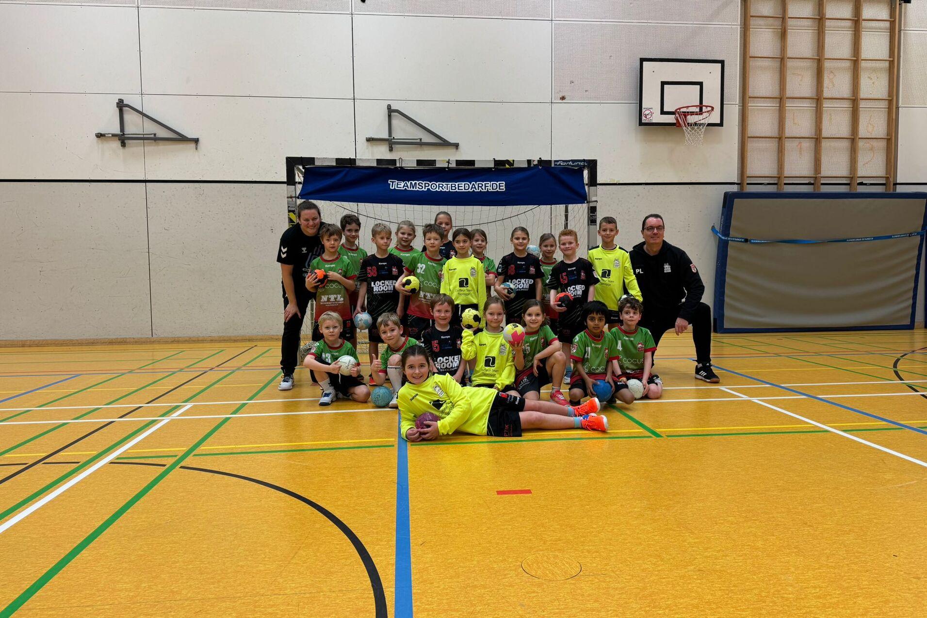 Gruppe von Kindern in Sportkleidung, die in einer Turnhalle vor einem Fußballtor posieren. Trainer stehen hinter ihnen.