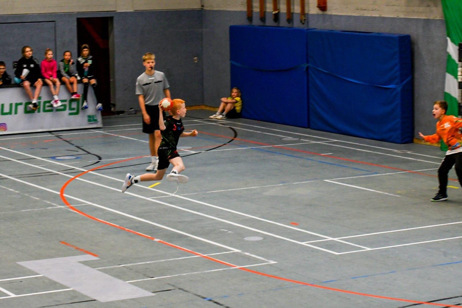 Ein junger Handballspieler springt zum Tor, w&auml;hrend der Torwart sich vorbereitet, im Hintergrund Zuschauer.