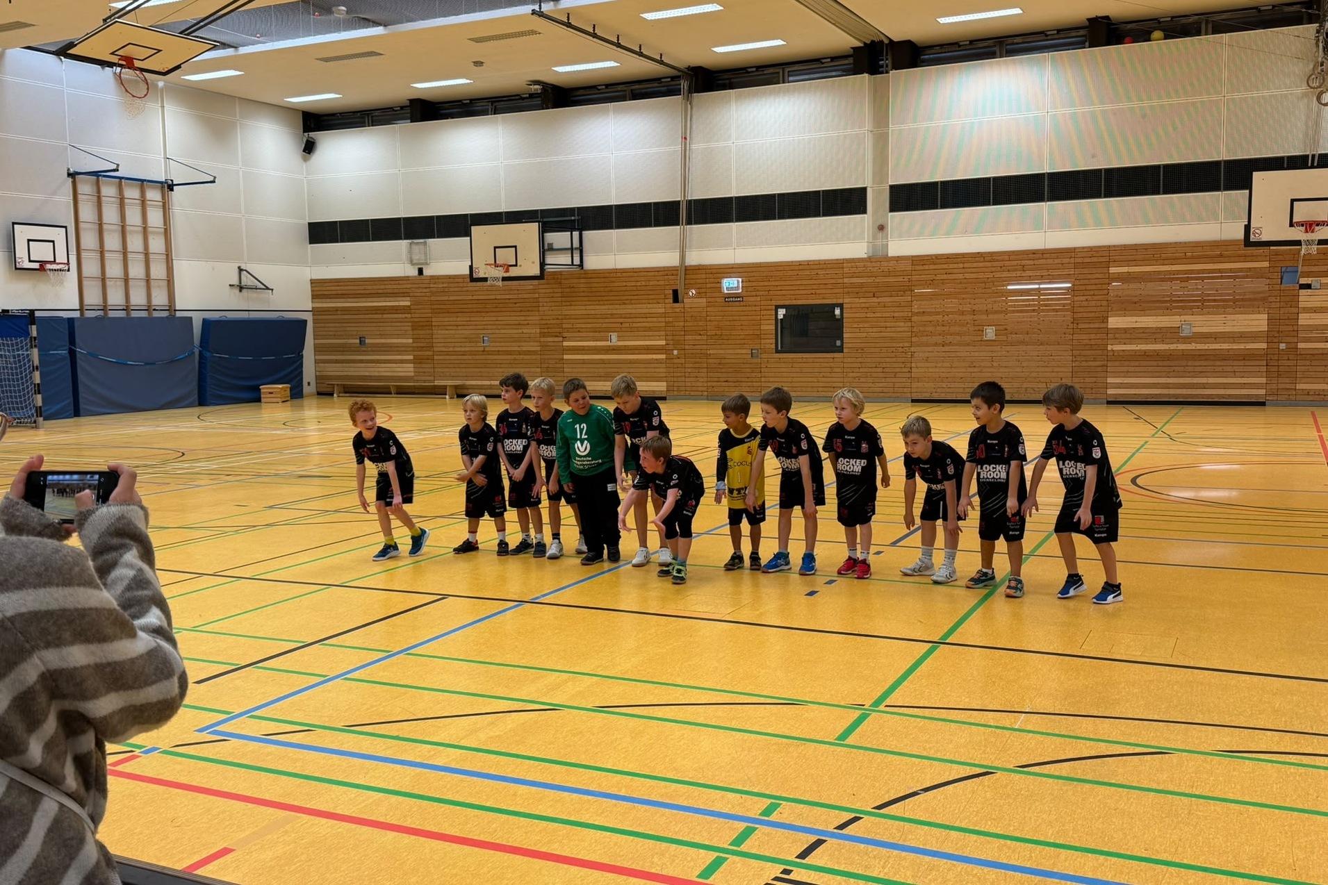 Gruppe von Kindern in Sportkleidung steht auf einer Turnhalle mit Linien und Basketballschultern im Hintergrund.