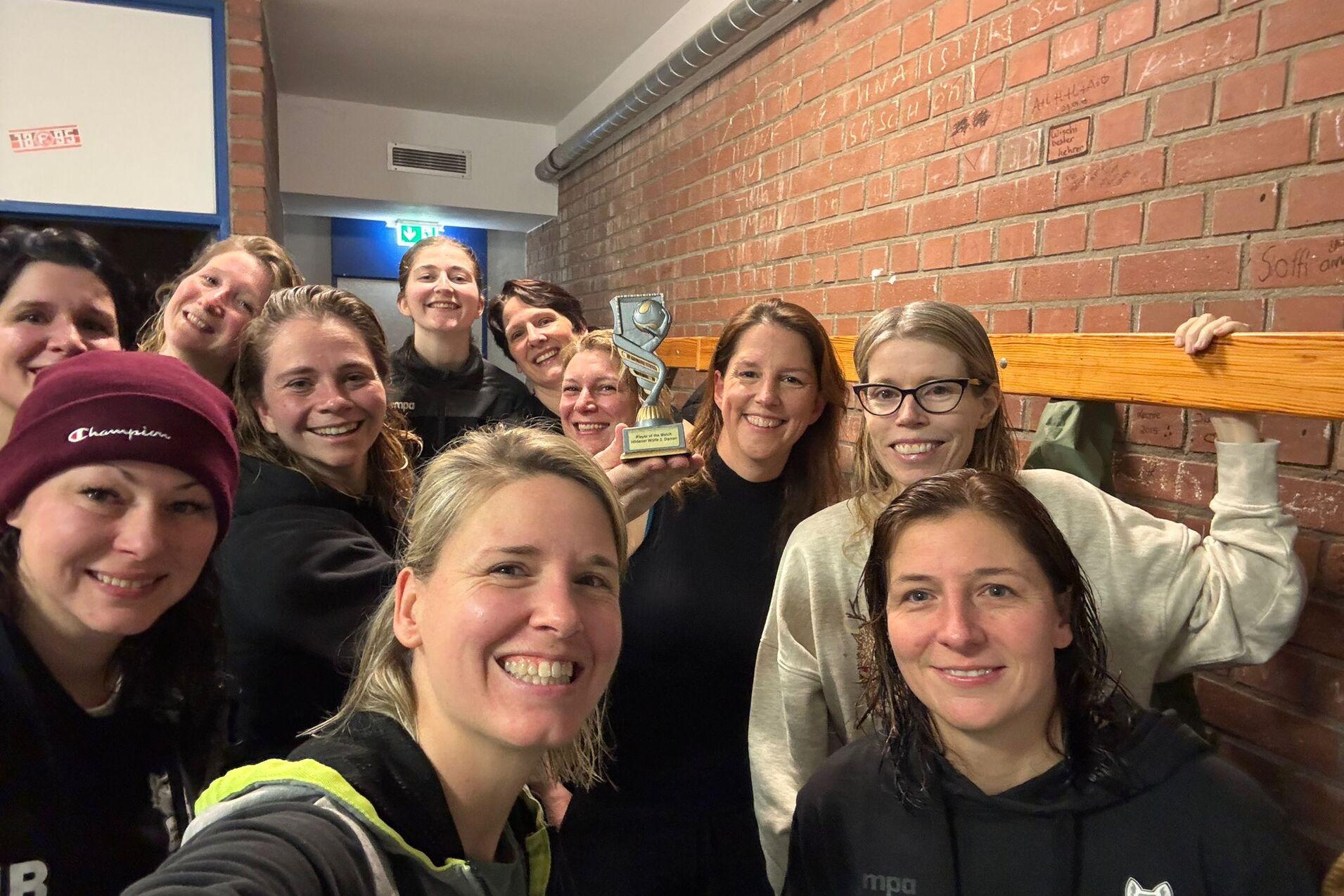 Gruppe von zehn Frauen posiert lächelnd für ein Selfie in einer sportlichen Umgebung mit einer Trophäe im Hintergrund.