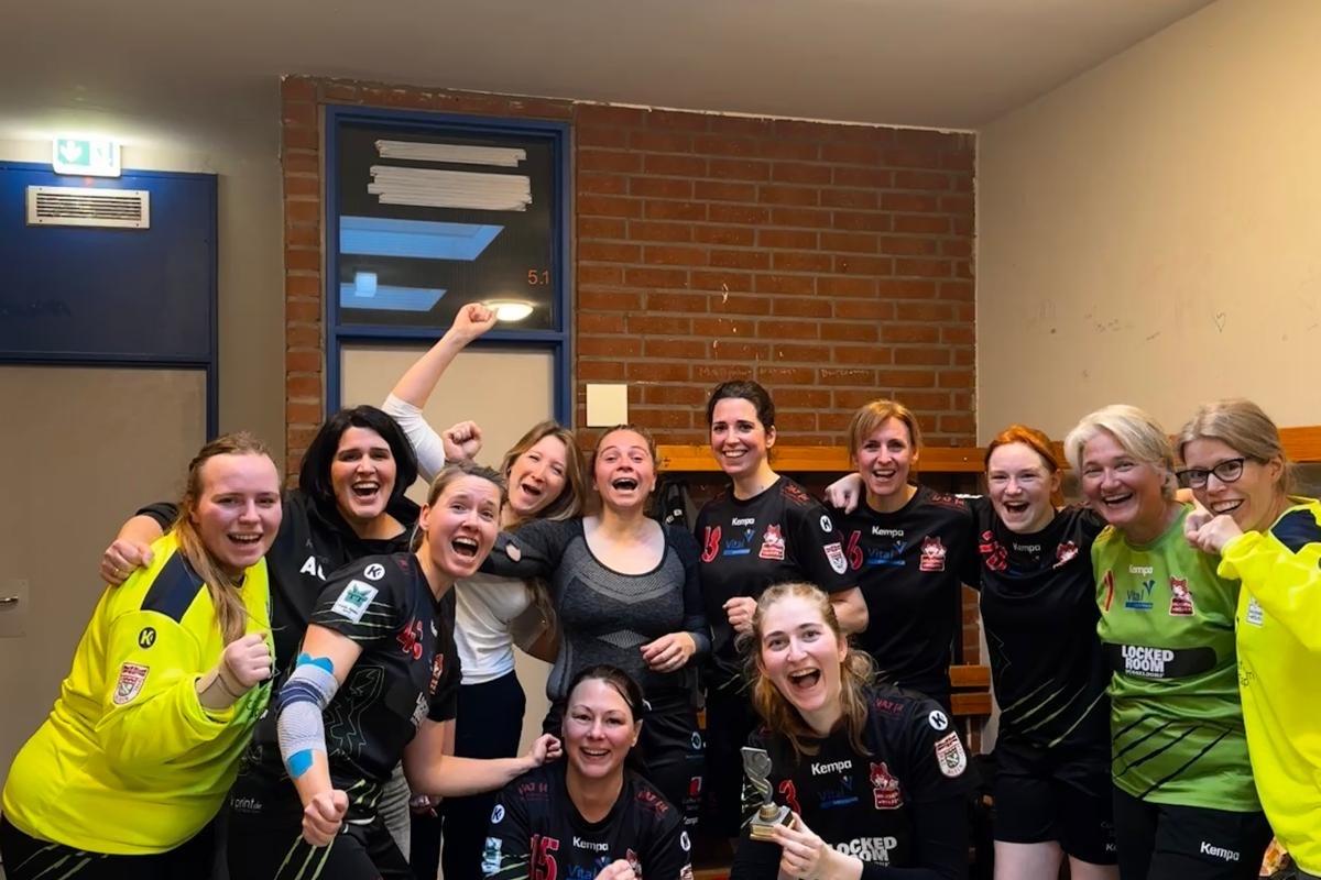 Gruppe von 13 jubelnden Frauen in Sportkleidung stehen freudig in einem Umkleideraum.