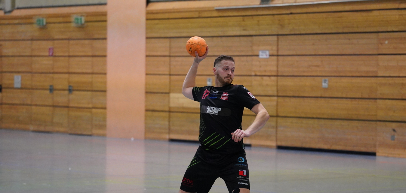Spieler in schwarzem Trikot wirft einen Handball in einer Sporthalle, ein weiterer Spieler im Hintergrund in gelbem Trikot.
