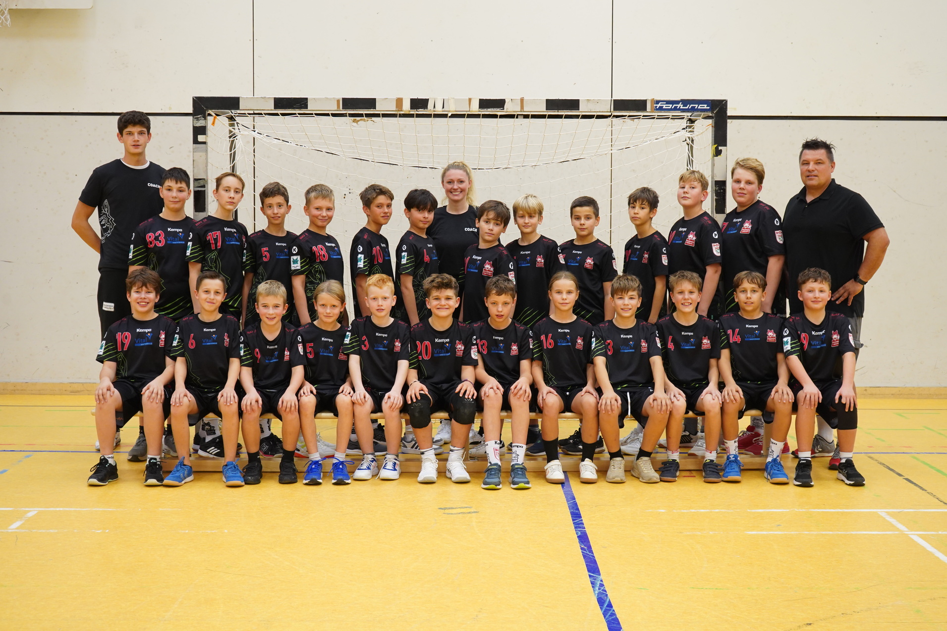 Gruppenfoto einer Jungen-Handballmannschaft in schwarzen Trikots vor einem Tor in einer Sporthalle.