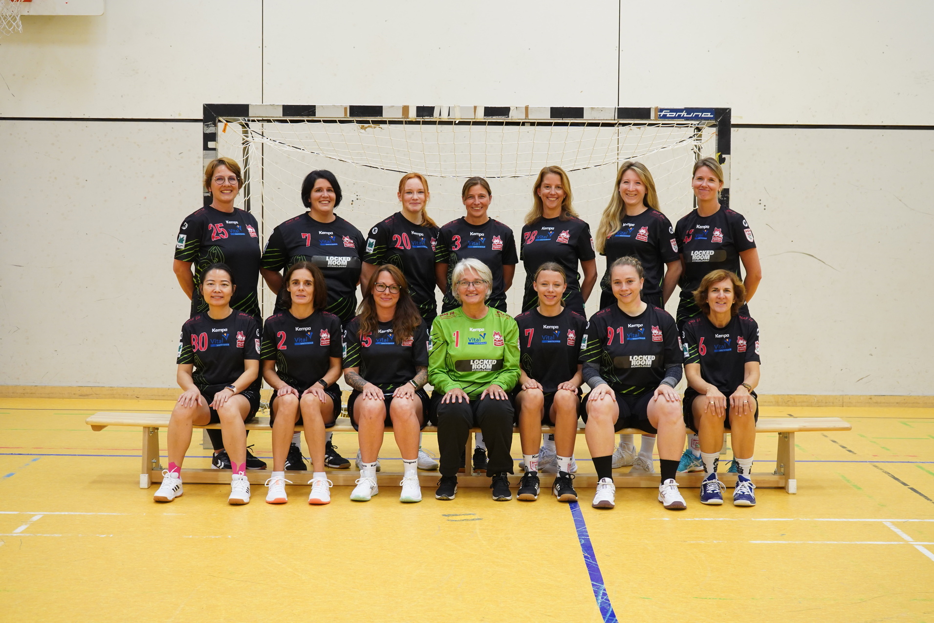 Gruppenfoto eines Handballteams mit 14 Spielerinnen und einer Trainerin in Sportkleidung vor einem Handballtor in einer Halle.