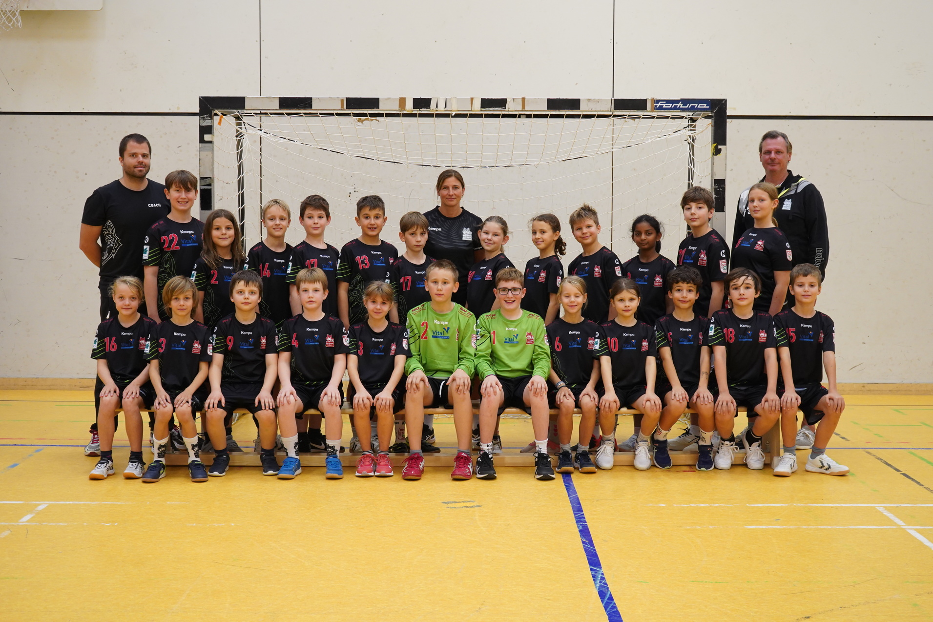 Gruppenfoto einer Handballmannschaft mit Trainer und Betreuern vor einem Tor in einer Sporthalle.