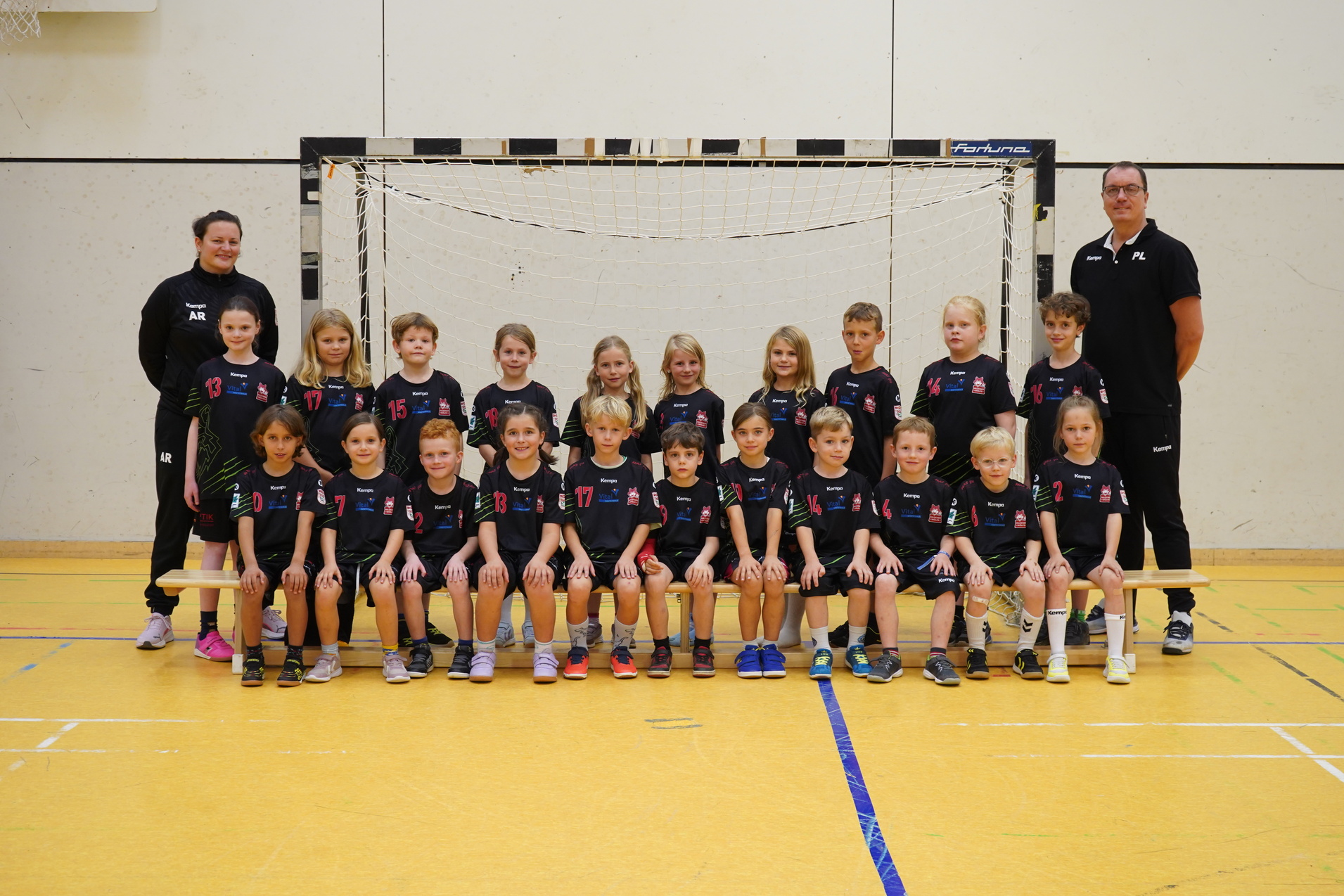 Gruppenbild einer Jugendmannschaft mit Trainer, alle in schwarzen Trikots vor einem Handballtor in einer Sporthalle.