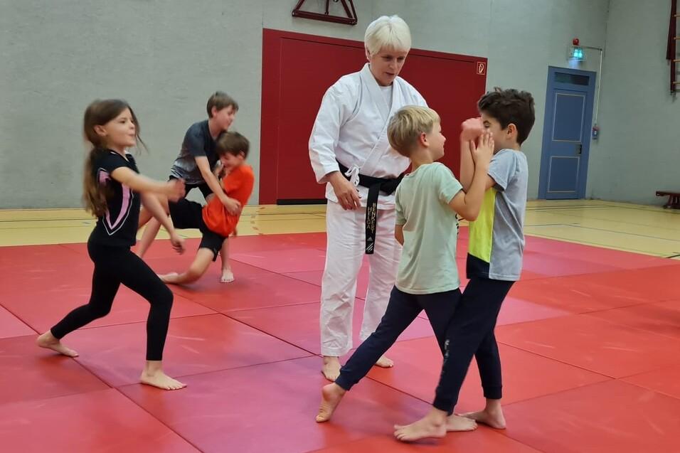 Kinder trainieren in einem Turnraum mit roten Matten, während ein Trainer Anweisungen gibt.