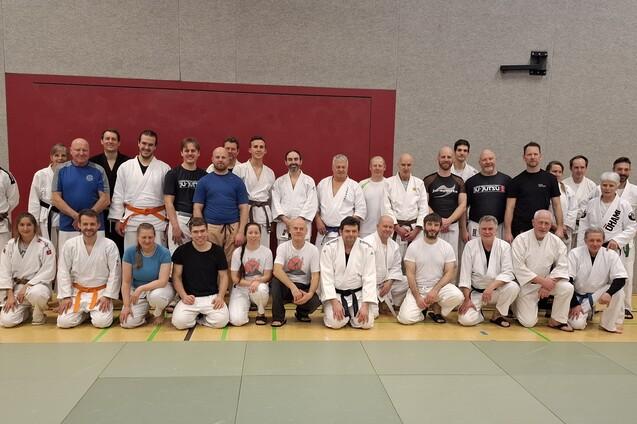 Gruppenfoto von Judo-Praktizierenden in wei&szlig;en Anz&uuml;gen auf einer Trainingsmatte, vor einer grauen Wand.