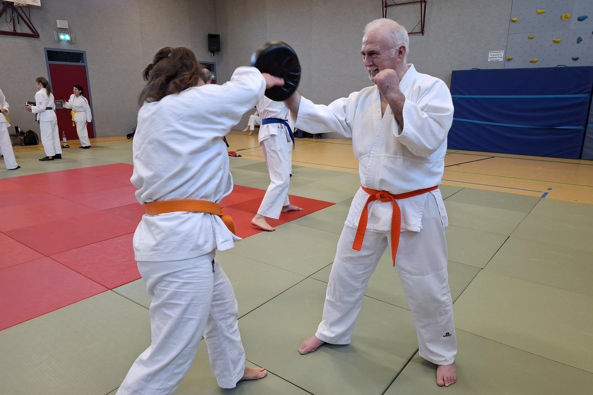 Zwei Karateka in wei&szlig;en Gi-Kleidung &uuml;ben Techniken auf einer Matte, ein Teilnehmer tr&auml;gt einen orangen G&uuml;rtel.