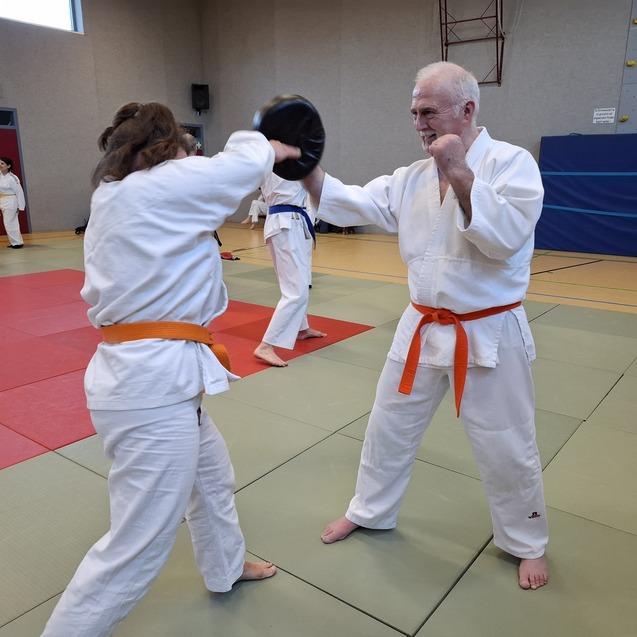 Zwei Karateka in wei&szlig;en Gi-Kleidung &uuml;ben Techniken auf einer Matte, ein Teilnehmer tr&auml;gt einen orangen G&uuml;rtel.