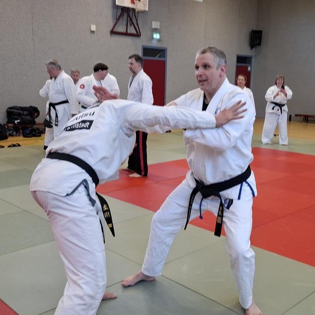 Zwei Kampfsportler in wei&szlig;en Gi-Kleidung &uuml;ben Techniken auf einer Trainingsmatte, w&auml;hrend weitere Teilnehmer im Hintergrund zuschauen.