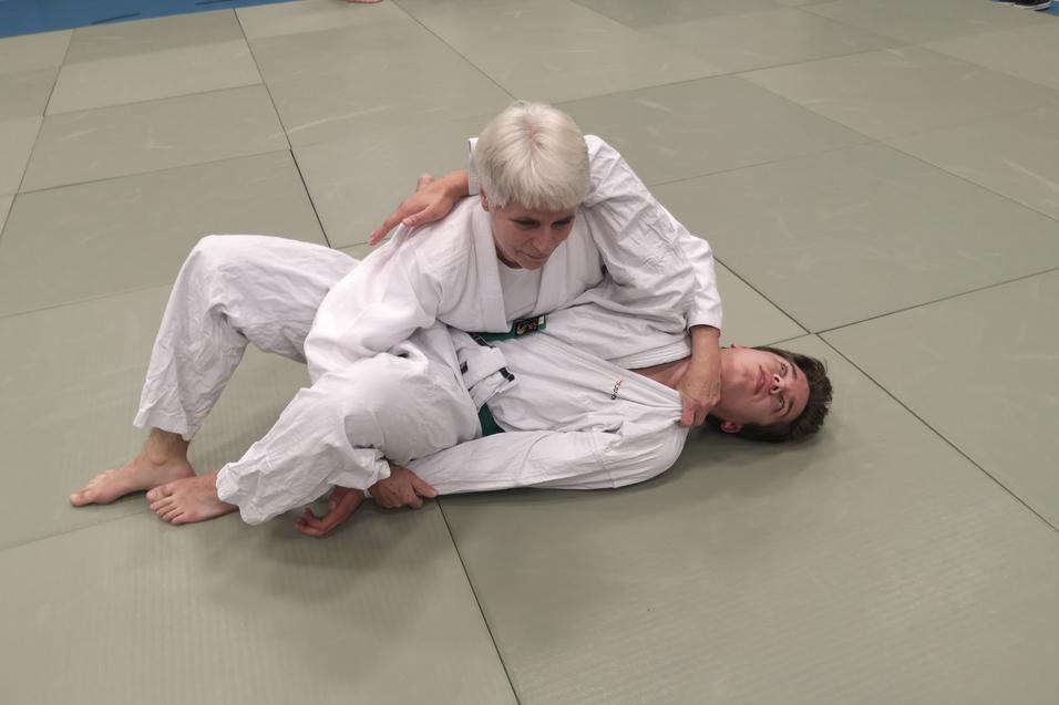 Zwei Judoka im weißen Gi üben einen Haltegriff auf einer Matte in einem Trainingsraum.
