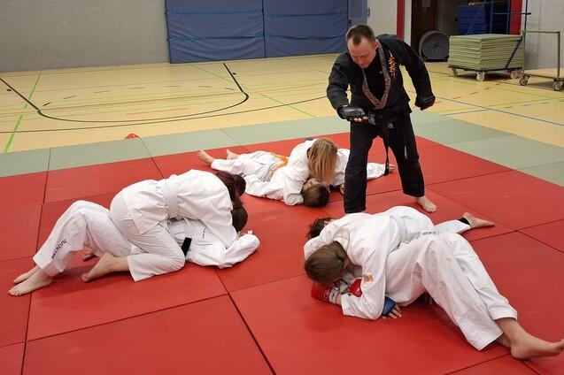 Trainer zeigt Techniken im Judo mit mehreren Sch&uuml;lern auf einer roten Matte in einem Sportraum.