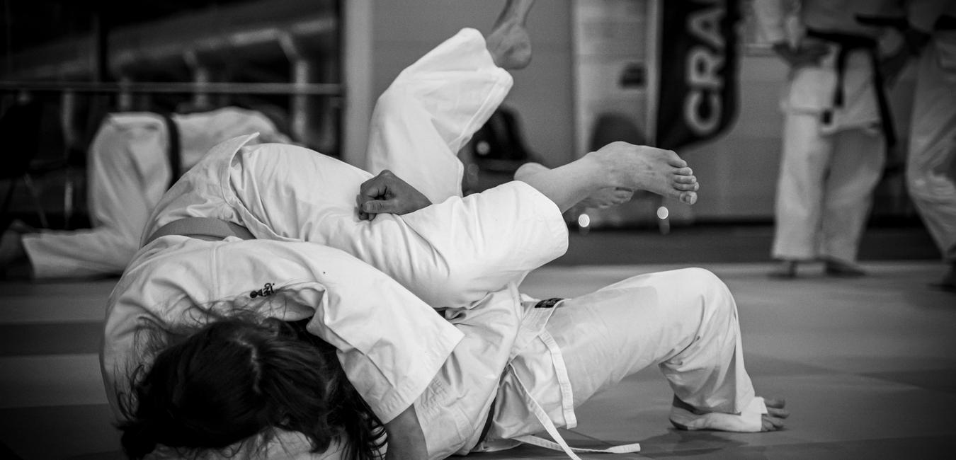 Judo-K&auml;mpfer in einer Kampfposition auf einer Matte, in traditioneller Judo-Uniform, im Hintergrund weitere Praktizierende.