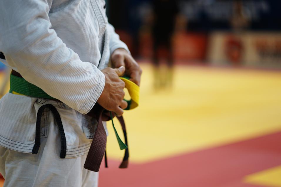 Nahaufnahme einer Person in einem wei&szlig;en Judogi, die bunte G&uuml;rtel in der Hand h&auml;lt, im Hintergrund eine Judomatte.