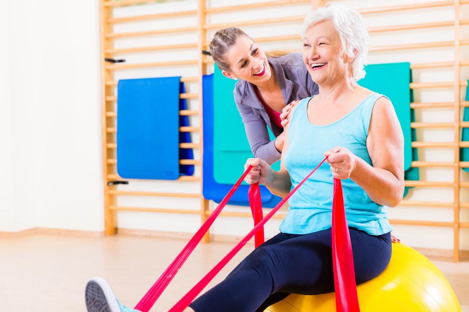 Junge Trainerin unterstützt lächelnde älterer Frau beim Training mit Widerstandsbändern auf einem Fitnessball.