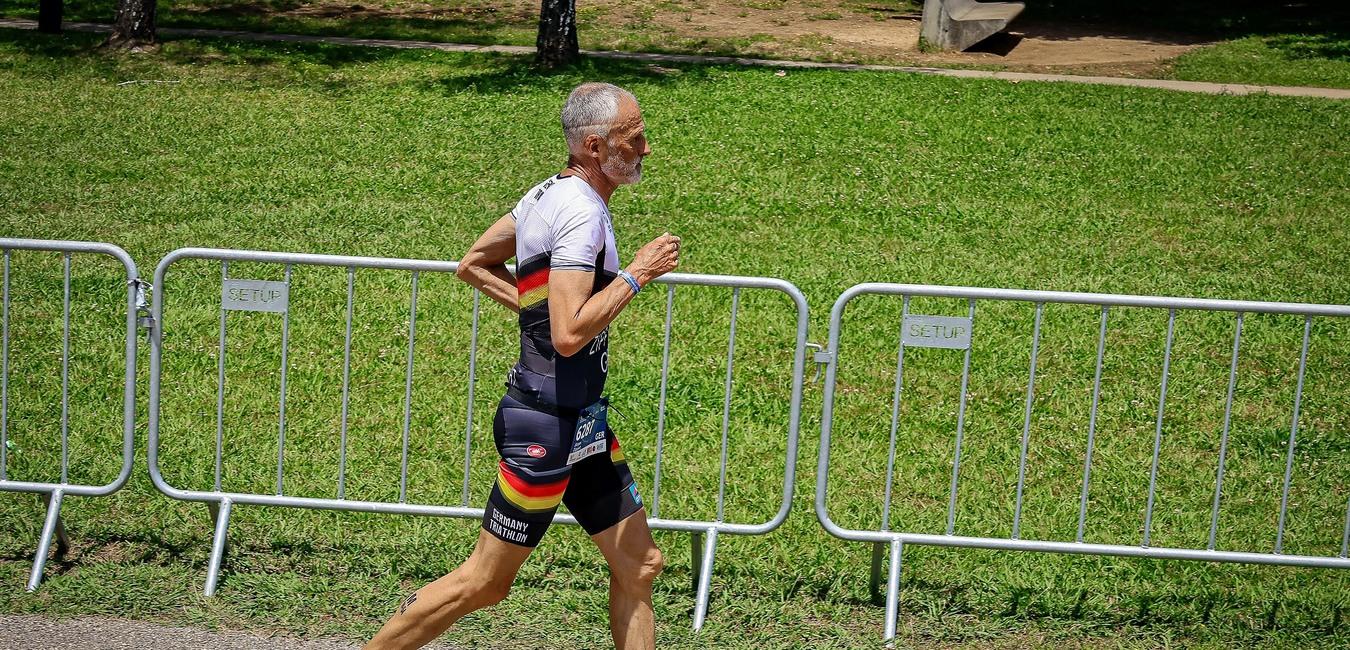 Älterer Mann in Sportkleidung läuft auf einem Weg, umgeben von Absperrgittern und grünem Rasen.