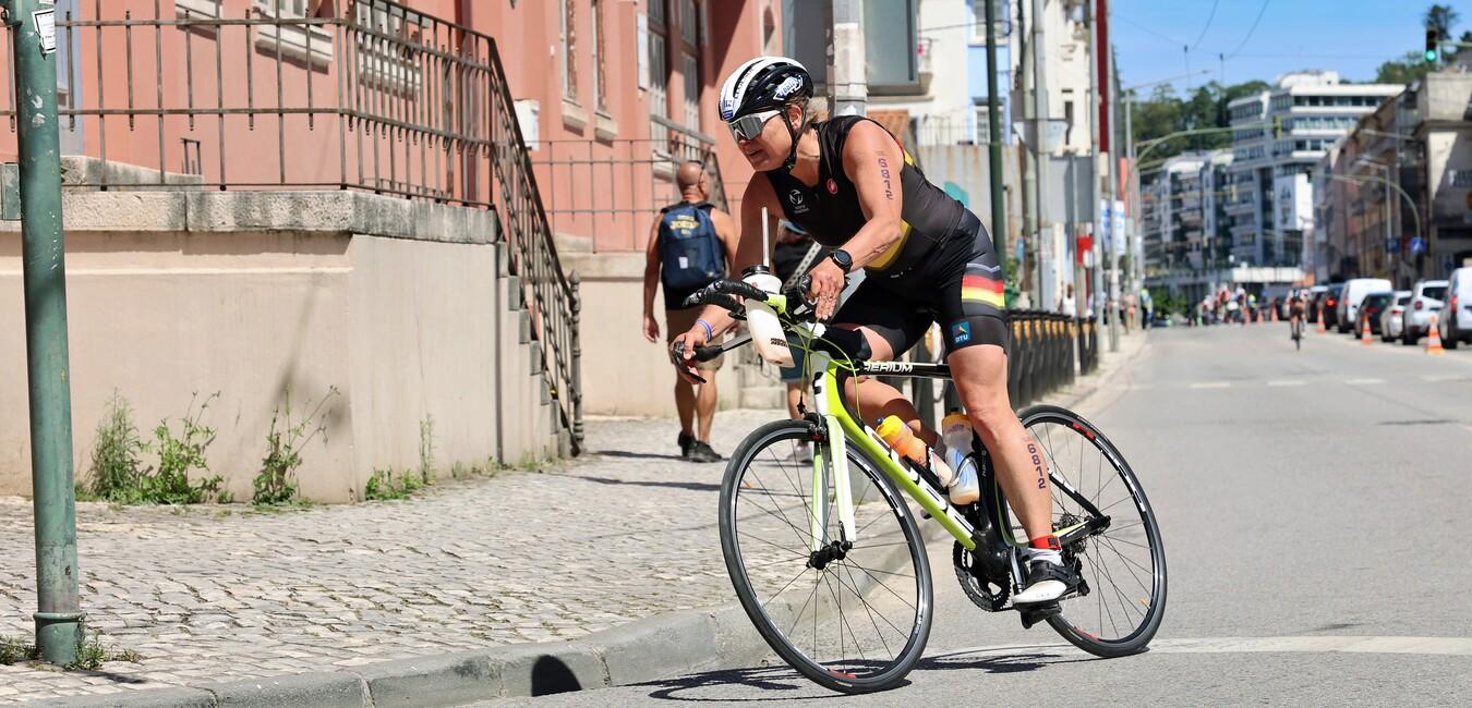 Sportler fährt auf einem Fahrrad die Straße entlang, mit farbenfrohem Trikot und Helm. Im Hintergrund ist städtische Architektur zu sehen.