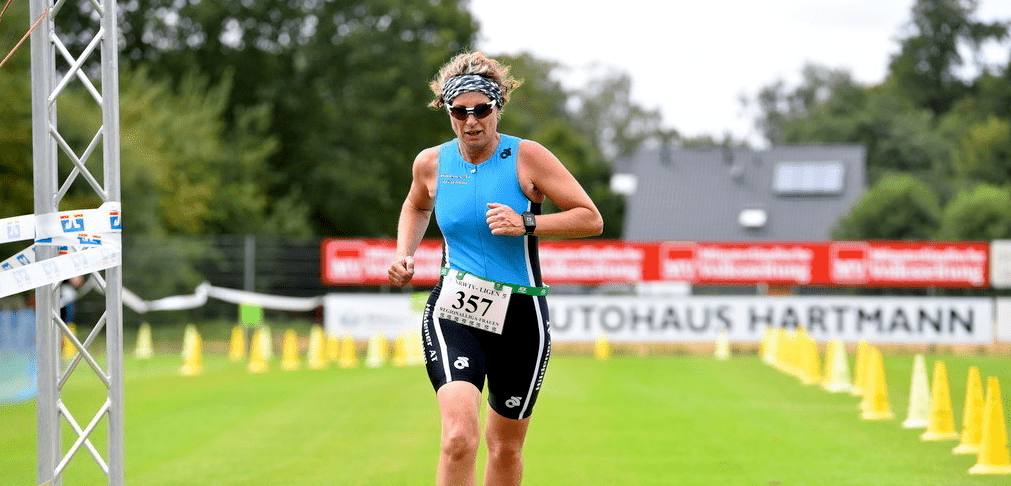 Sportlerin in blauer Wettkleidung überquert die Ziellinie eines Wettbewerbs auf einer grünen Rasenfläche.