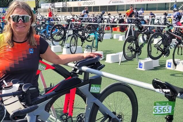 Frau mit Sonnenbrille steht neben ihrem Rennrad in einer Wechselzone beim Triathlon, mit weiteren Fahrr&auml;dern im Hintergrund.