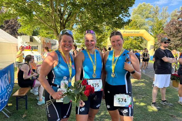 Drei Frauen in Sportkleidung halten Medaillen und Blumen, stehen l&auml;chelnd vor einer gr&uuml;nen Kulisse nach einem Wettkampf.