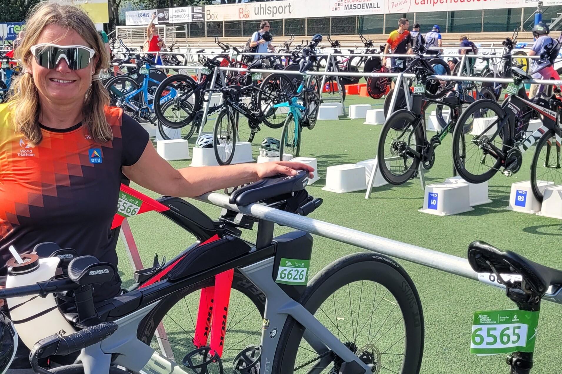 Frau mit Sonnenbrille steht neben ihrem Rennrad in einer Wechselzone beim Triathlon, mit weiteren Fahrr&auml;dern im Hintergrund.