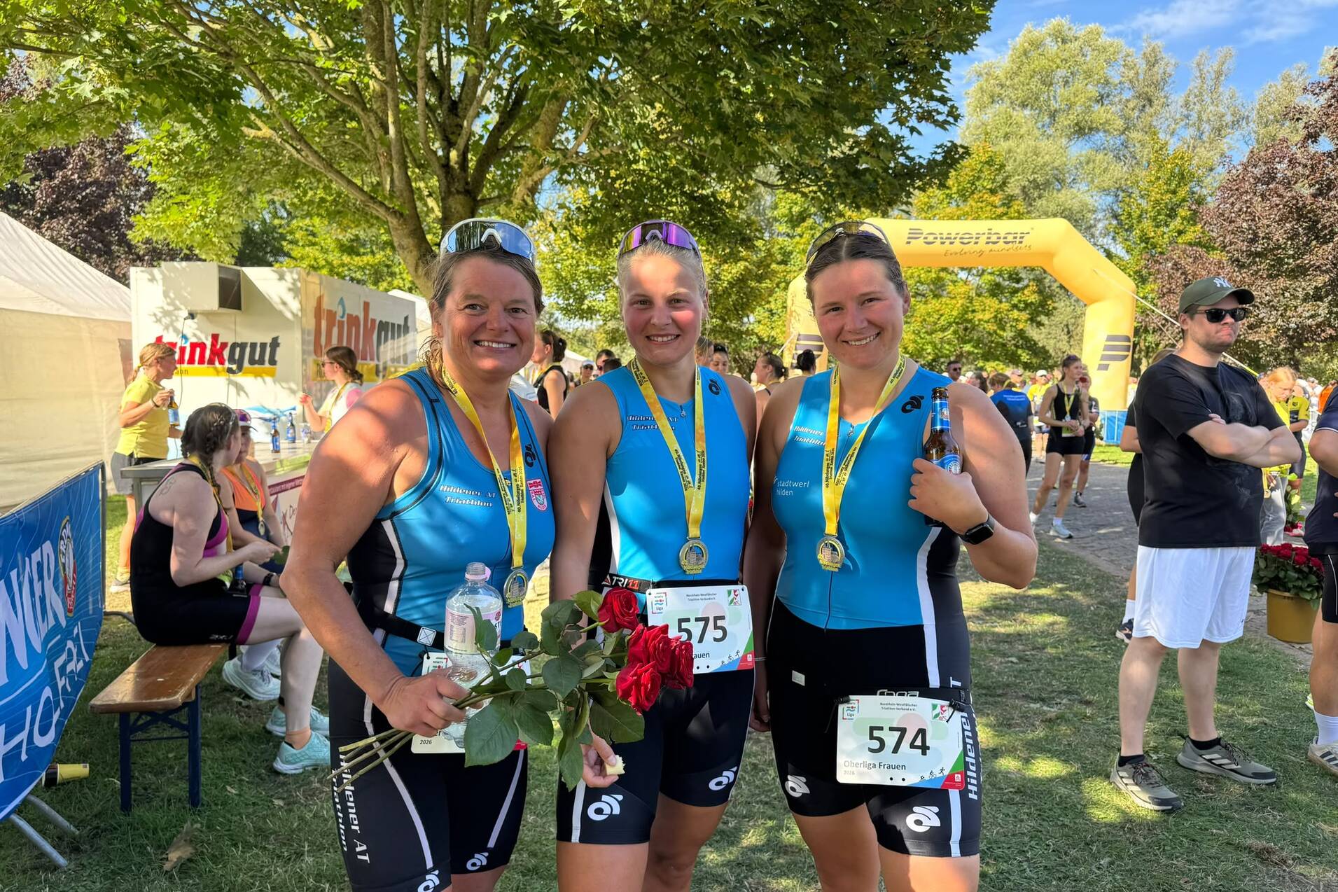 Drei Frauen in Sportkleidung halten Medaillen und Blumen, stehen lächelnd vor einer grünen Kulisse nach einem Wettkampf.