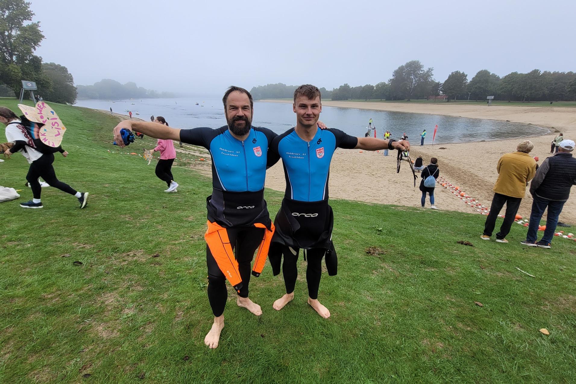 Zwei Männer in Neoprenanzügen stehen mit ausgebreiteten Armen am Ufer eines Badestrandes, im Hintergrund sind Menschen im Wasser.