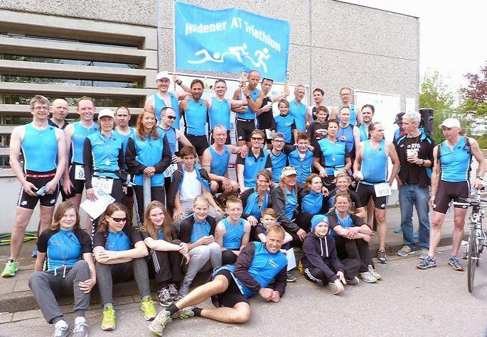 Gruppenzusammenkunft von Triathleten in blauen Wettkampfanzügen vor einem Banner des Hildener Triathlons.