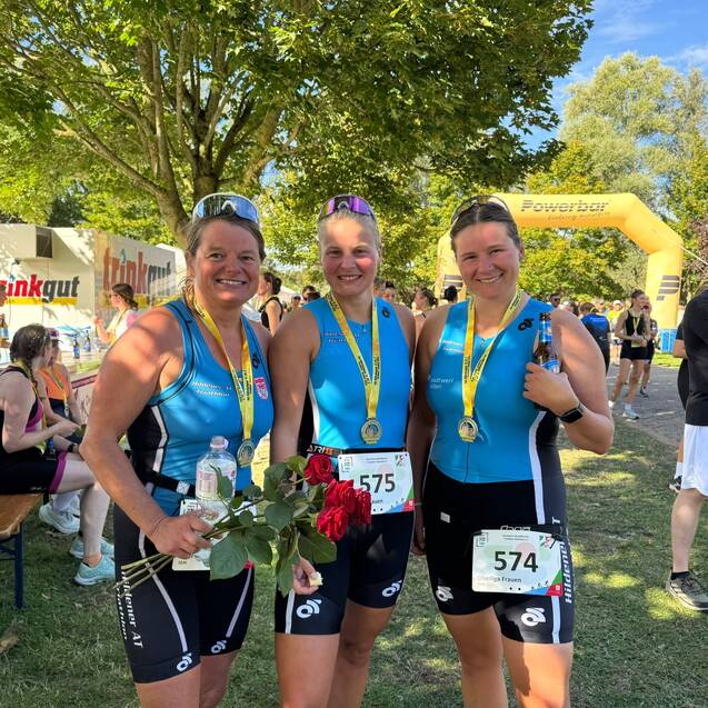 Drei Frauen in Sportkleidung halten Medaillen und Blumen, stehen lächelnd vor einer grünen Kulisse nach einem Wettkampf.